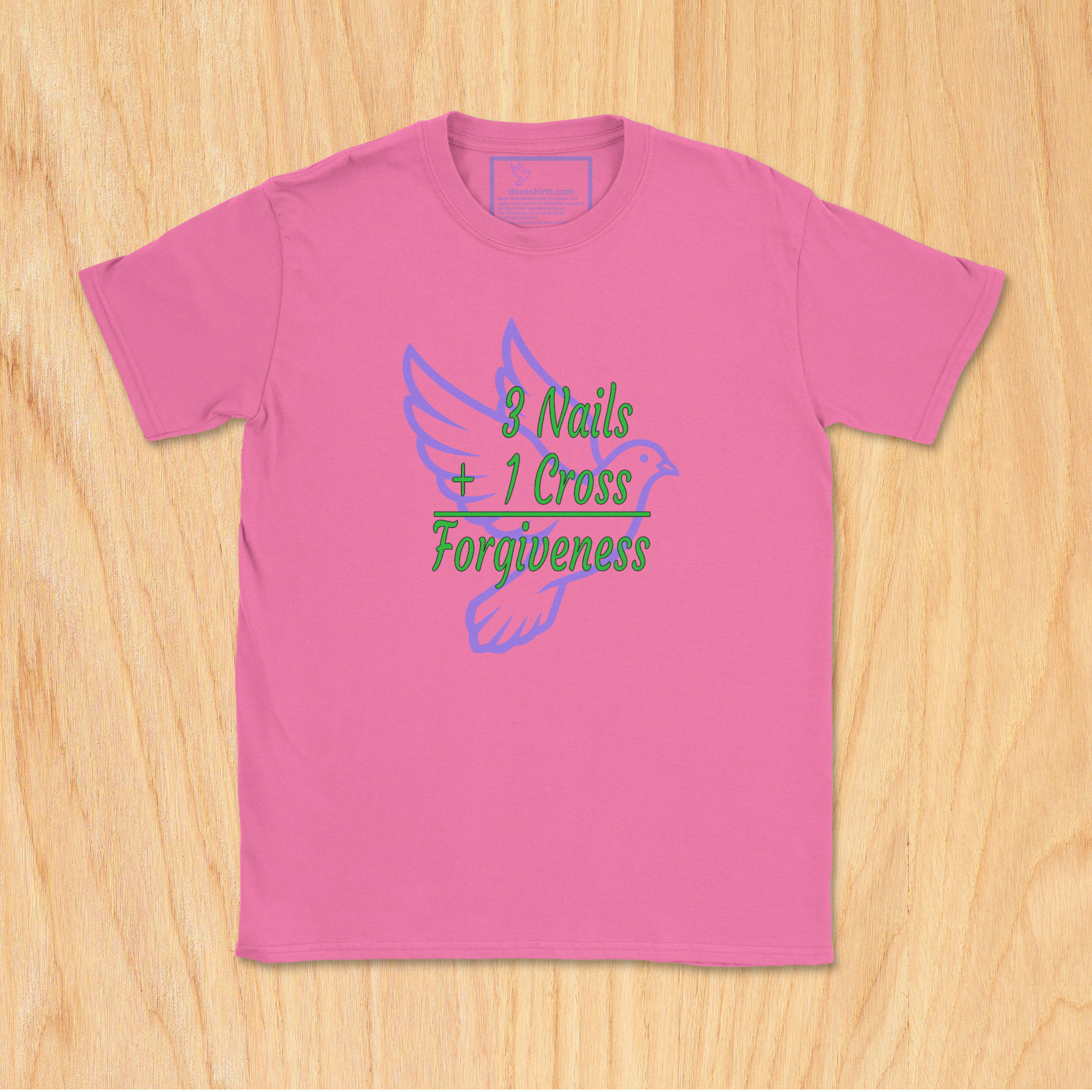 A azalea pink short-sleeve t-shirt with a graphic on the front that reads '8 Nails + 1 Cross Forgiveness' in green and purple text over a purple dove illustration, laid flat on a light wood background.