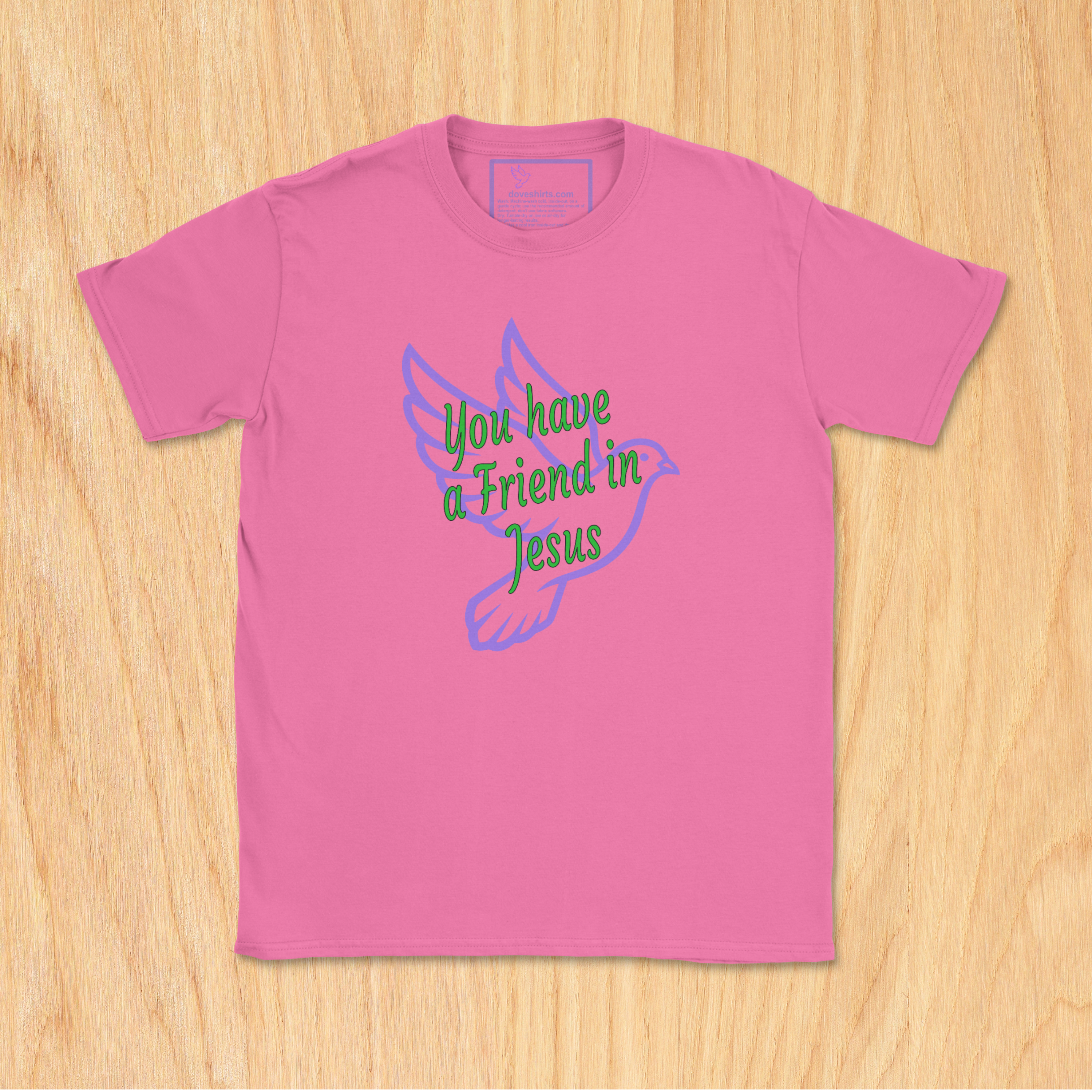 A azalea pink short-sleeve t-shirt with a graphic on the front showing a purple dove and the text 'You have a Friend in Jesus' in green and purple, laid flat on a light wood background.