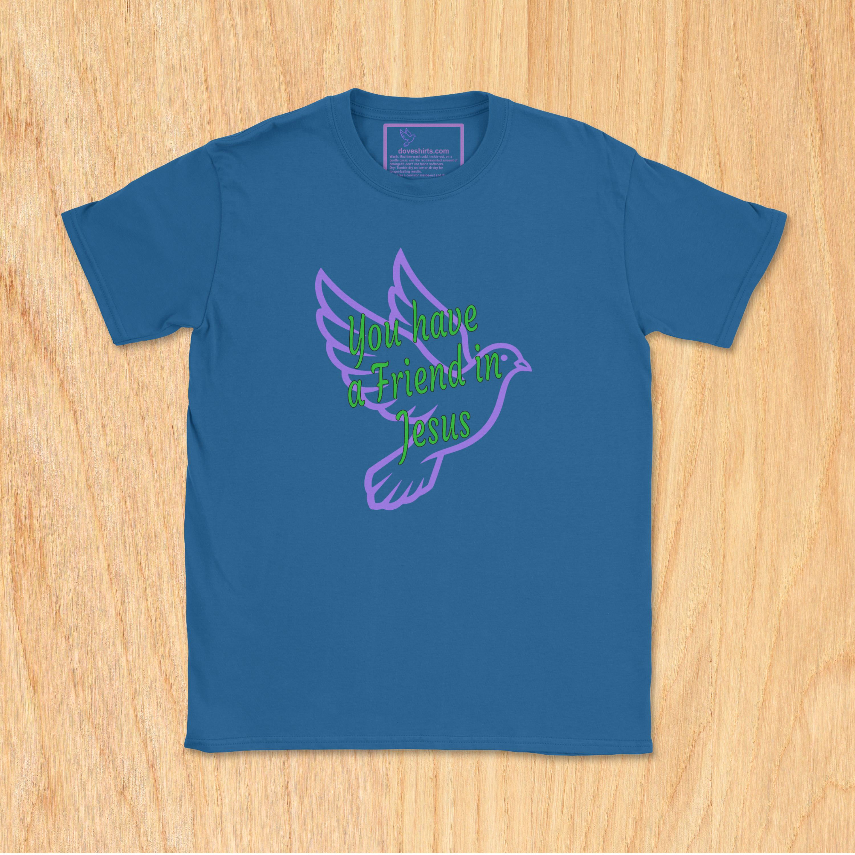 A royal short-sleeve t-shirt laid flat on a light wood background, featuring a purple outline of a dove and green cursive text that reads 'you have a friend in Jesus' across the chest.
