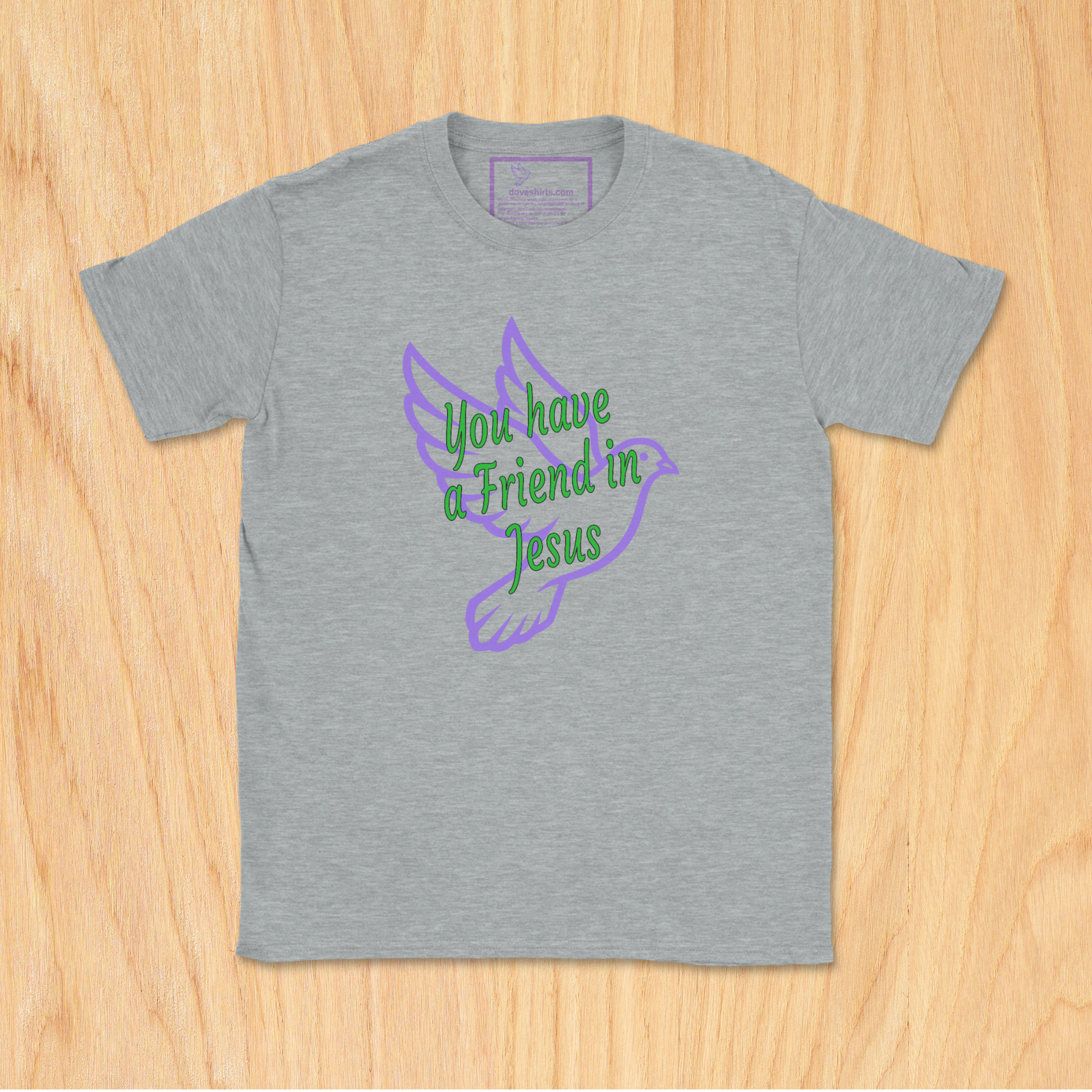 A heather sport gray short-sleeve t-shirt laid flat on a light wood background. The shirt features a graphic of a purple dove with green text that reads 'You have a Friend in Jesus'.